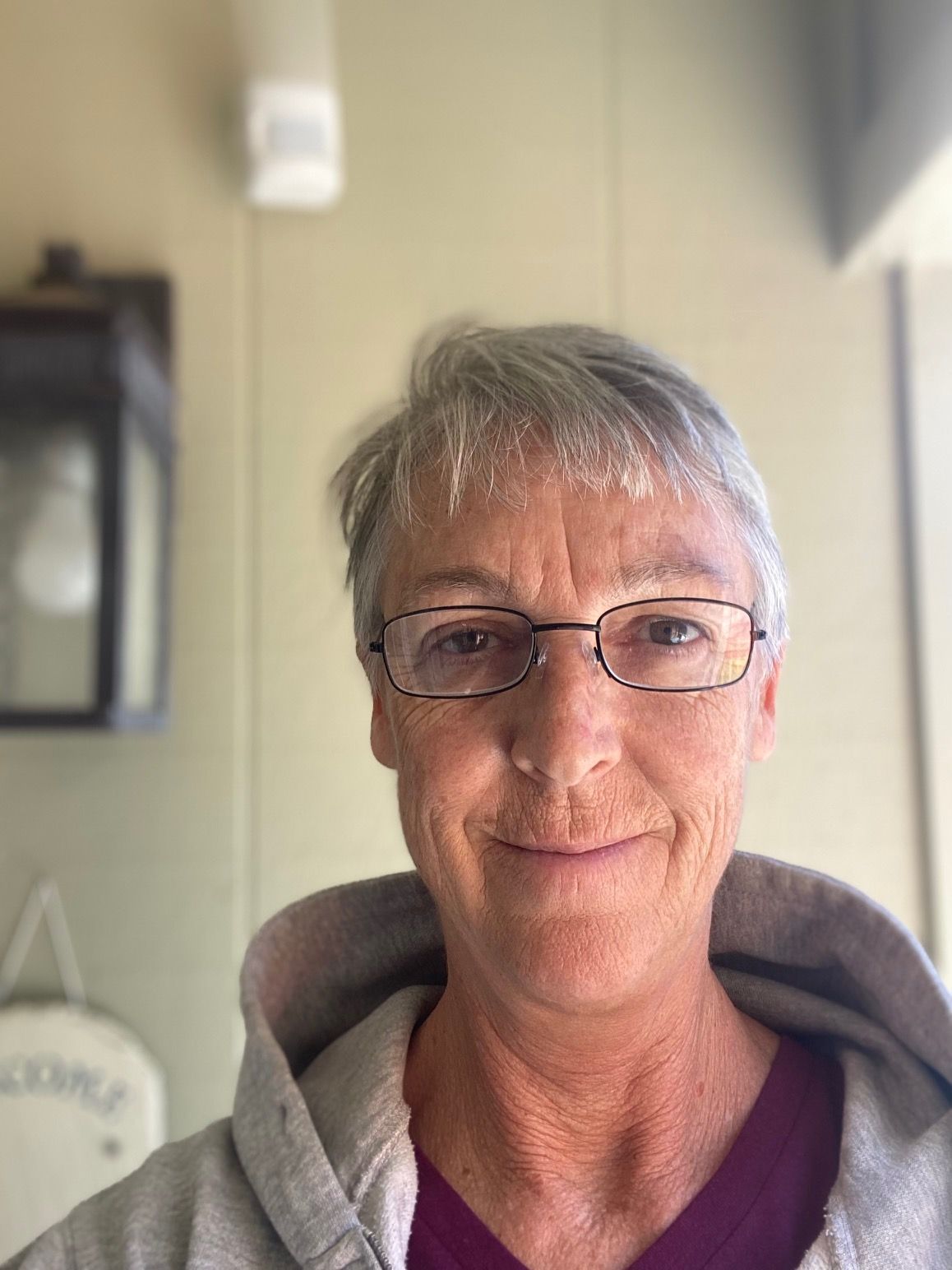 Woman with gray hair and glasses, smiling, wearing a hooded sweatshirt, outdoors.