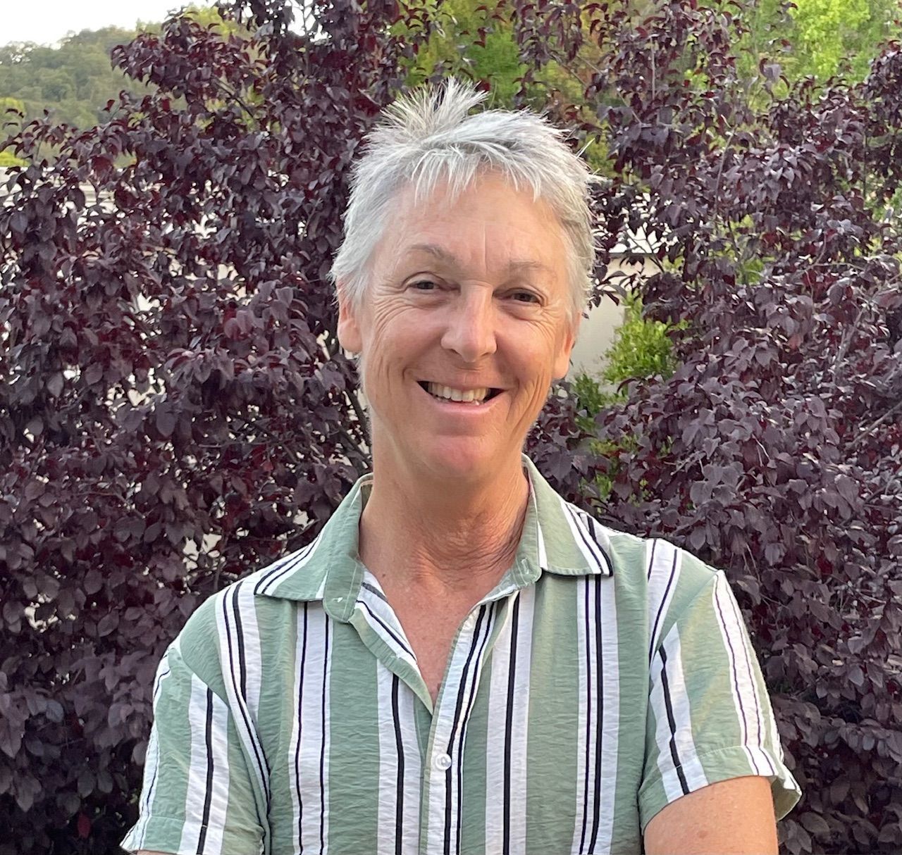 Woman with short gray hair, smiling, in green striped shirt; red bush background.