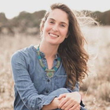 Juliana Sloane. Woman with long brown hair smiling wearing a blue top and sitting with her arms folded over her knees and sitting in a golden field.