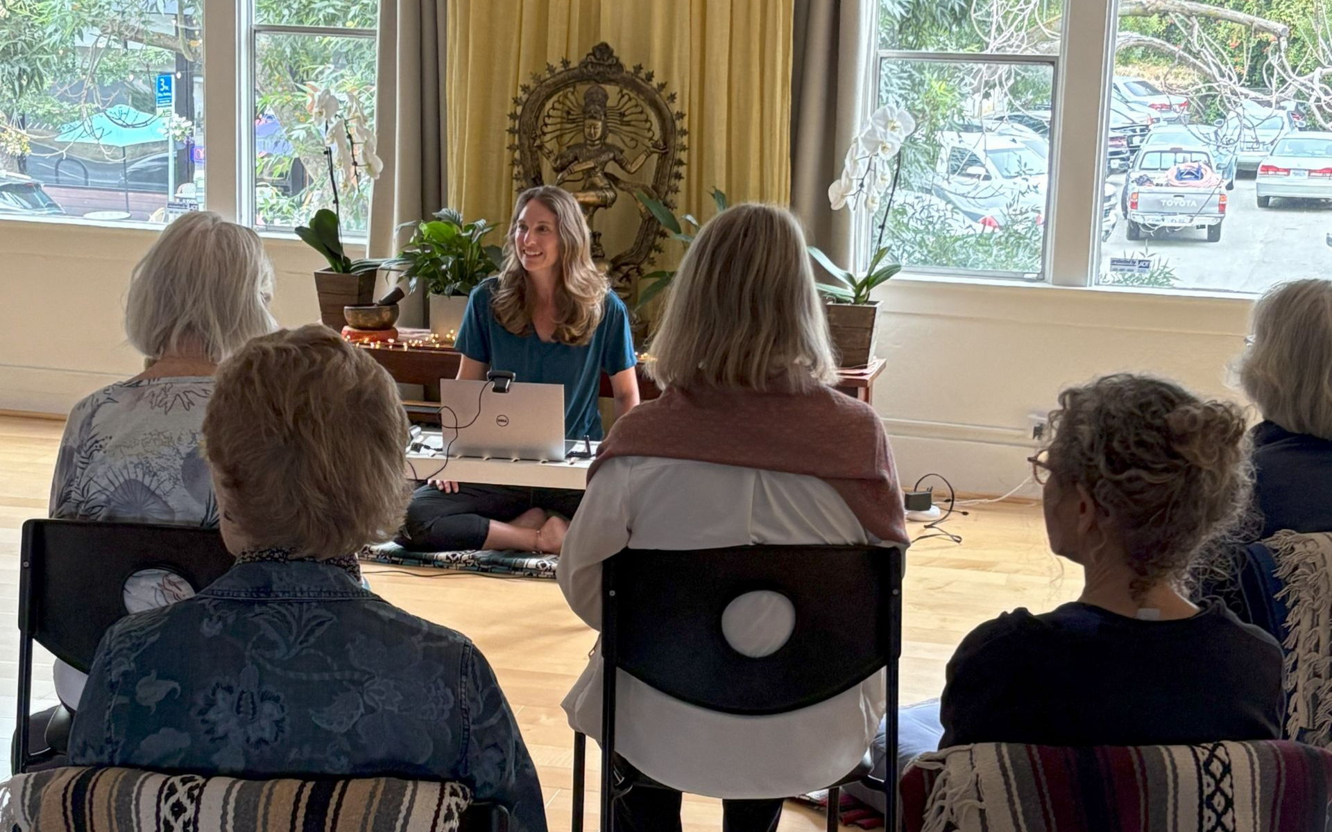 A woman leads a wellness class, facing attendees seated in chairs. Indoor setting.