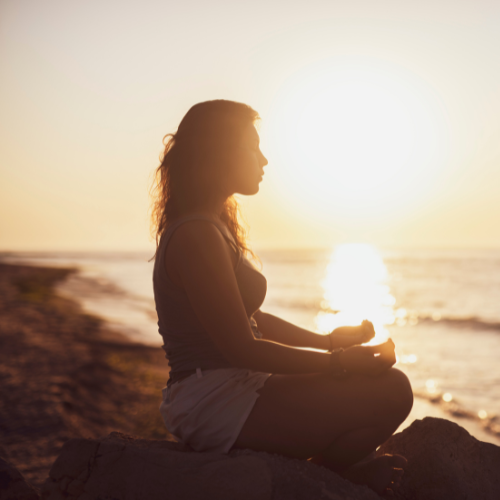 Woman meditating at sunset on a beach, hands in lap, silhouetted against the sun and ocean.