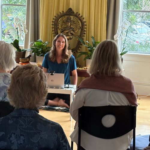 Woman leading a yoga class, seated in front of students, in a sunny room with decor.