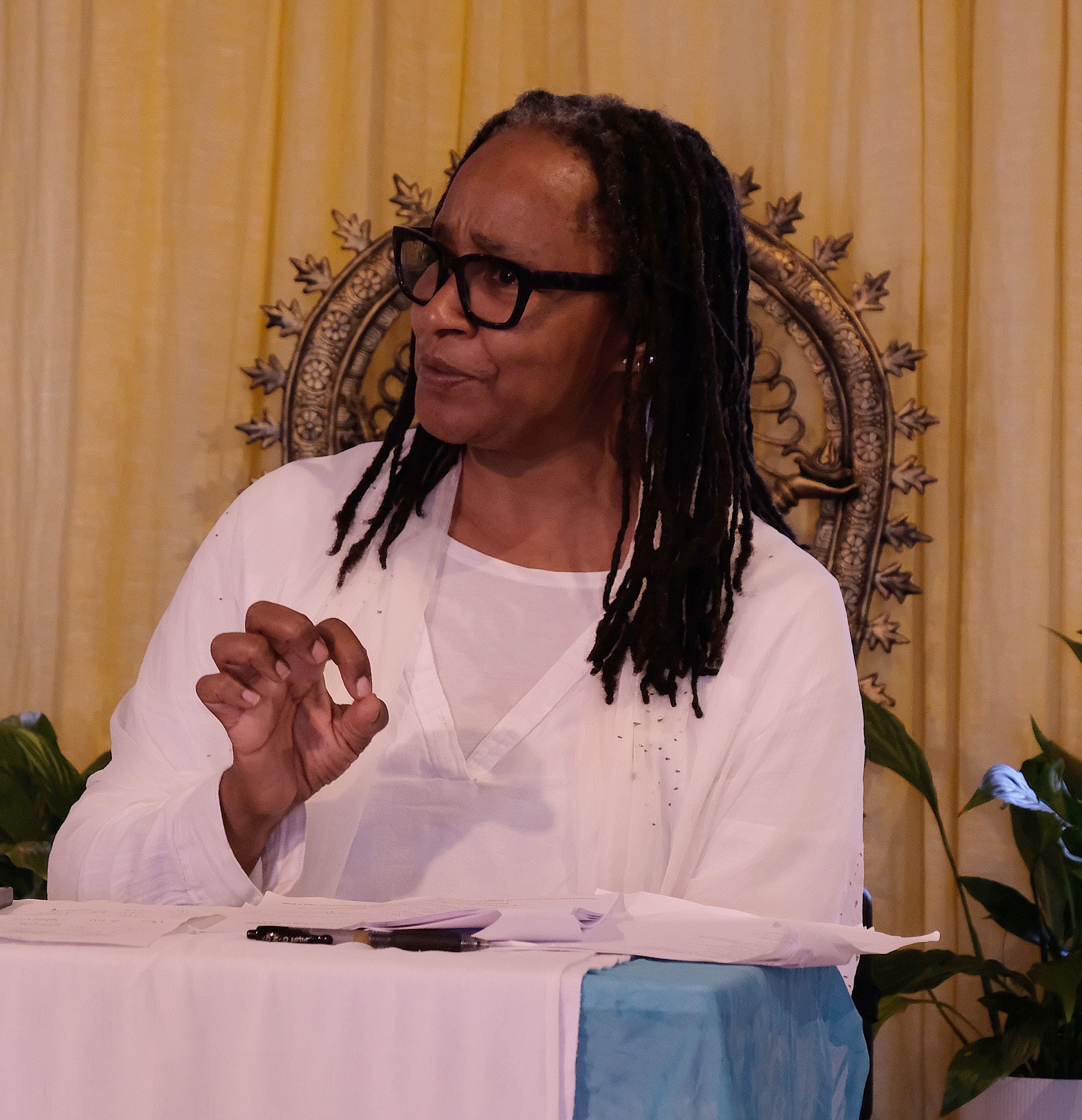 Woman with glasses, dreadlocks, and white shirt gestures while speaking at a table; setting is light and airy.