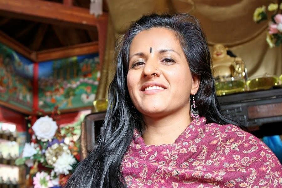 Woman with dark hair and bindi smiles, wearing a paisley shawl. Seated in temple, Buddha statue behind.