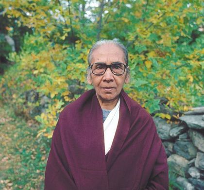 Elderly woman wearing glasses and burgundy shawl, standing outdoors near autumn foliage.