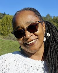 Woman with sunglasses smiles outdoors, wearing a white sweater, long dark dreadlocks, and earrings.