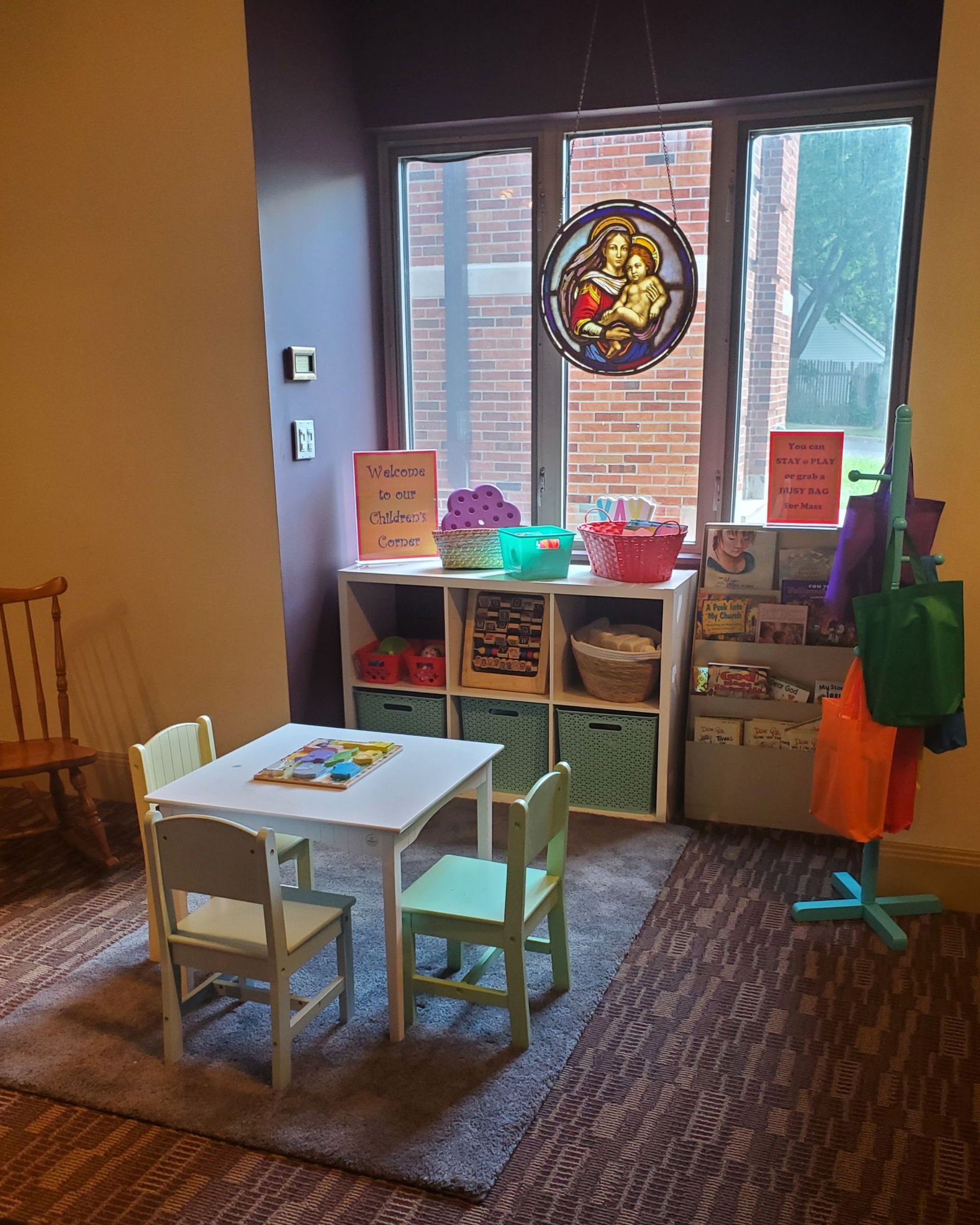 Children's Corner in the Gathering Space at Christ the King, featuring a wooden rocking chair, child-sized table and chairs, colorful busy bags, books, toys, and a stained-glass image of Mary holding the child Jesus.