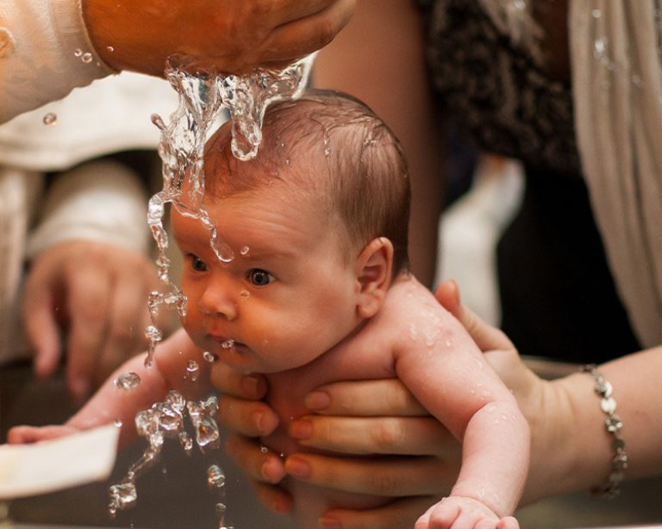 Holy water being poured out of a priest's hand to baptize an infant with their eyes open and held out by their mother's hands.