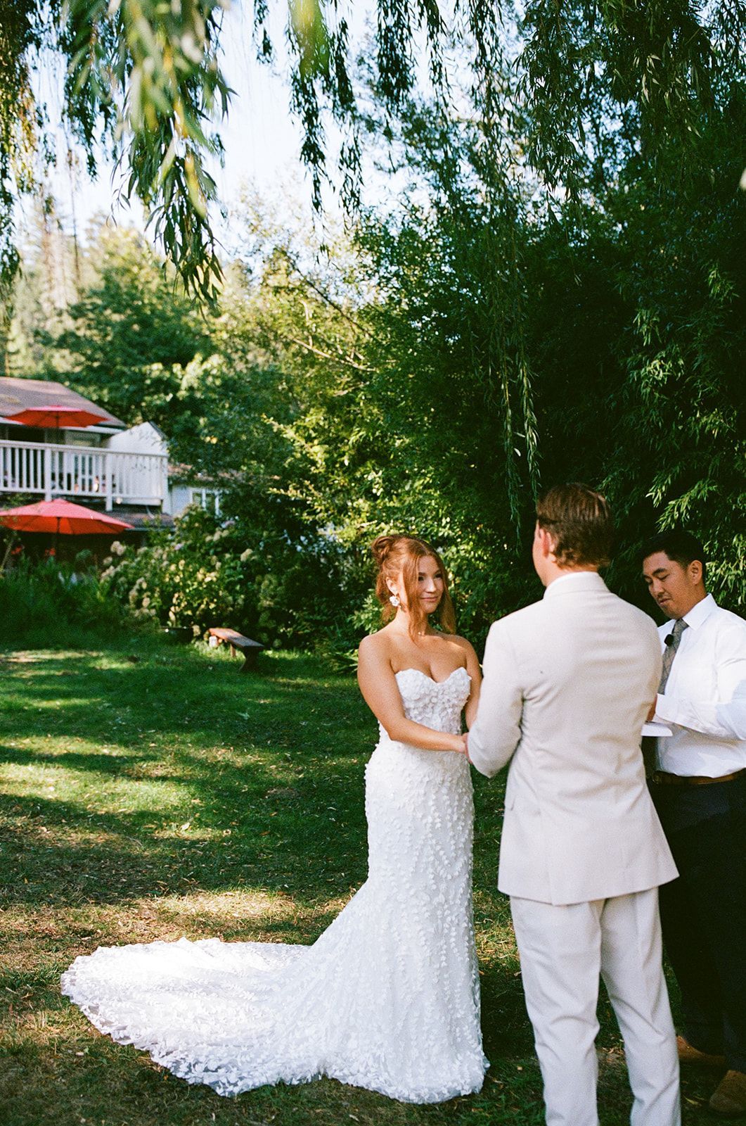 Bride and groom at outdoor wedding ceremony. Bride in white gown, groom in tan suit, officiant with them.