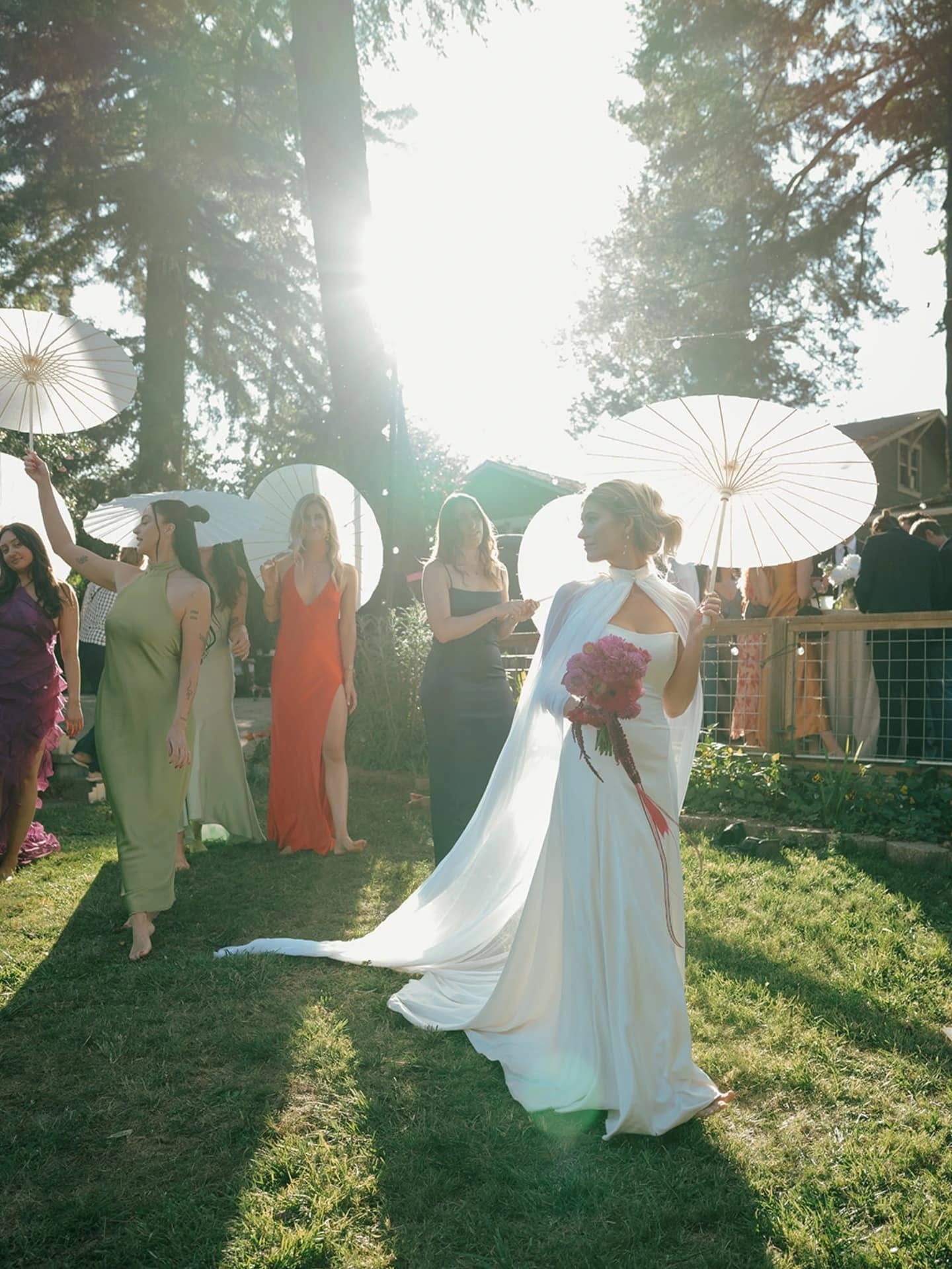 Bride in white gown with cape, carrying bouquet, walking toward camera on grass, sunlit with bridesmaids.
