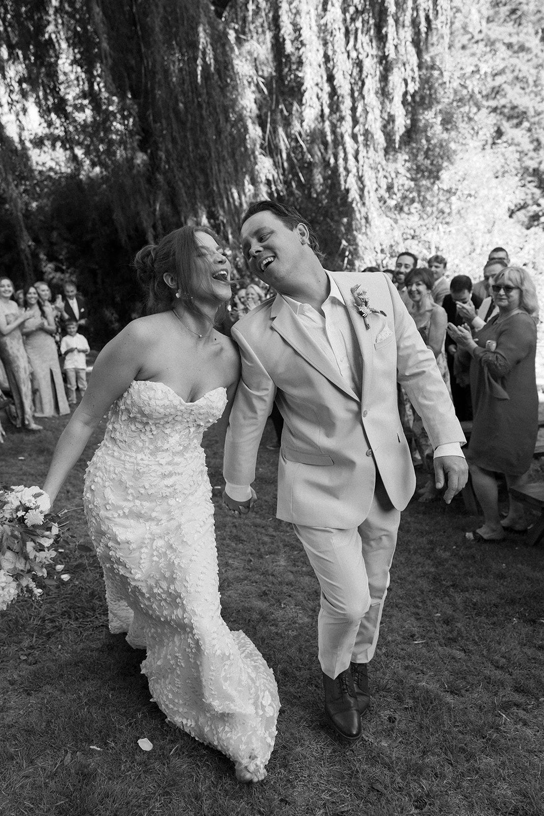 Newlyweds laughing, walking hand-in-hand after ceremony; bride in white gown, groom in suit; surrounded by applauding guests.