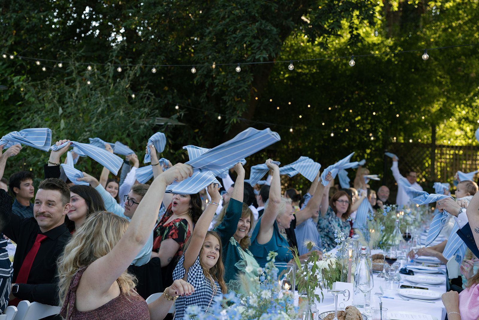People celebrating at an outdoor event, waving blue napkins above long table. String lights in background.