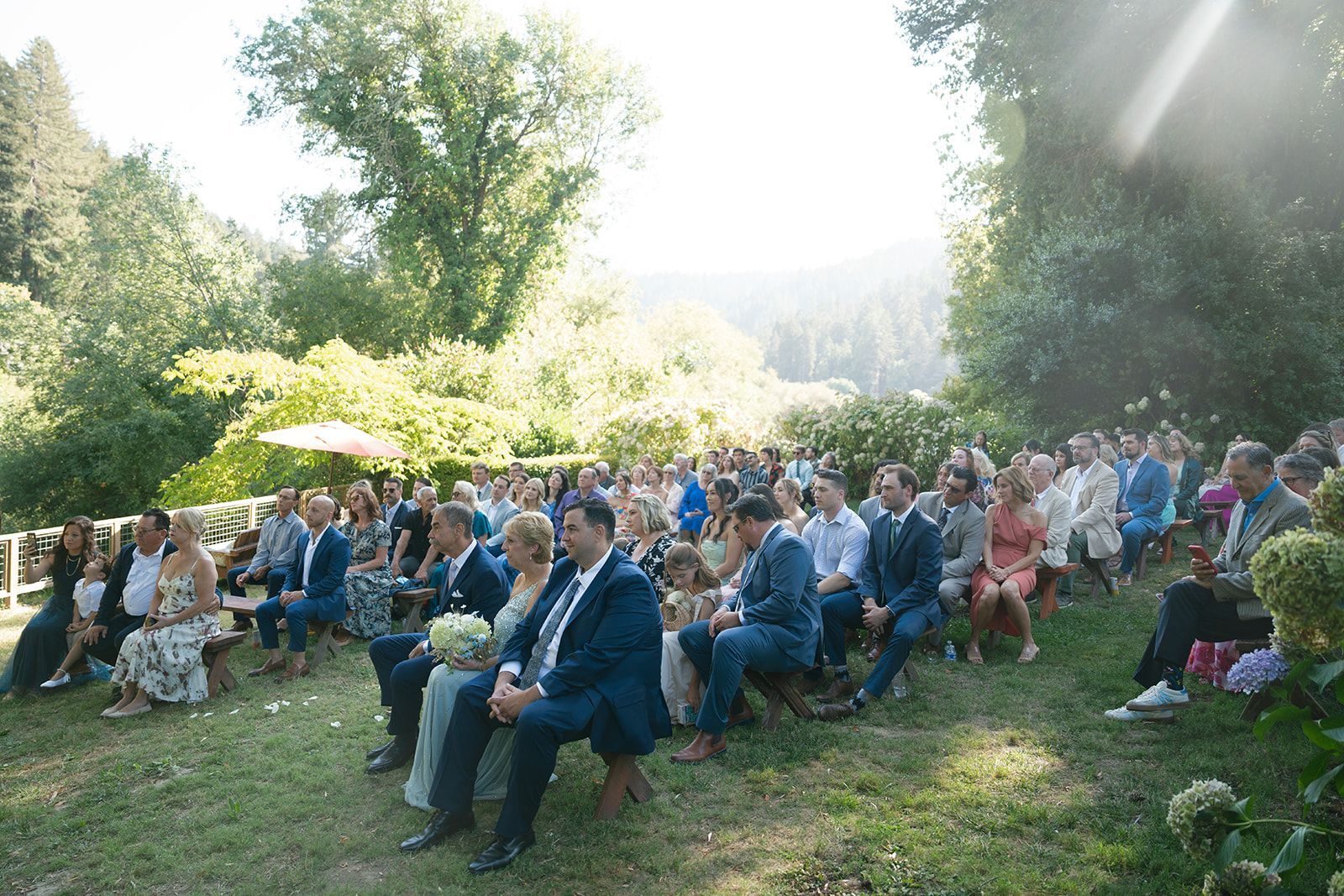 Wedding ceremony outdoors; guests seated on wooden benches, greenery in background.