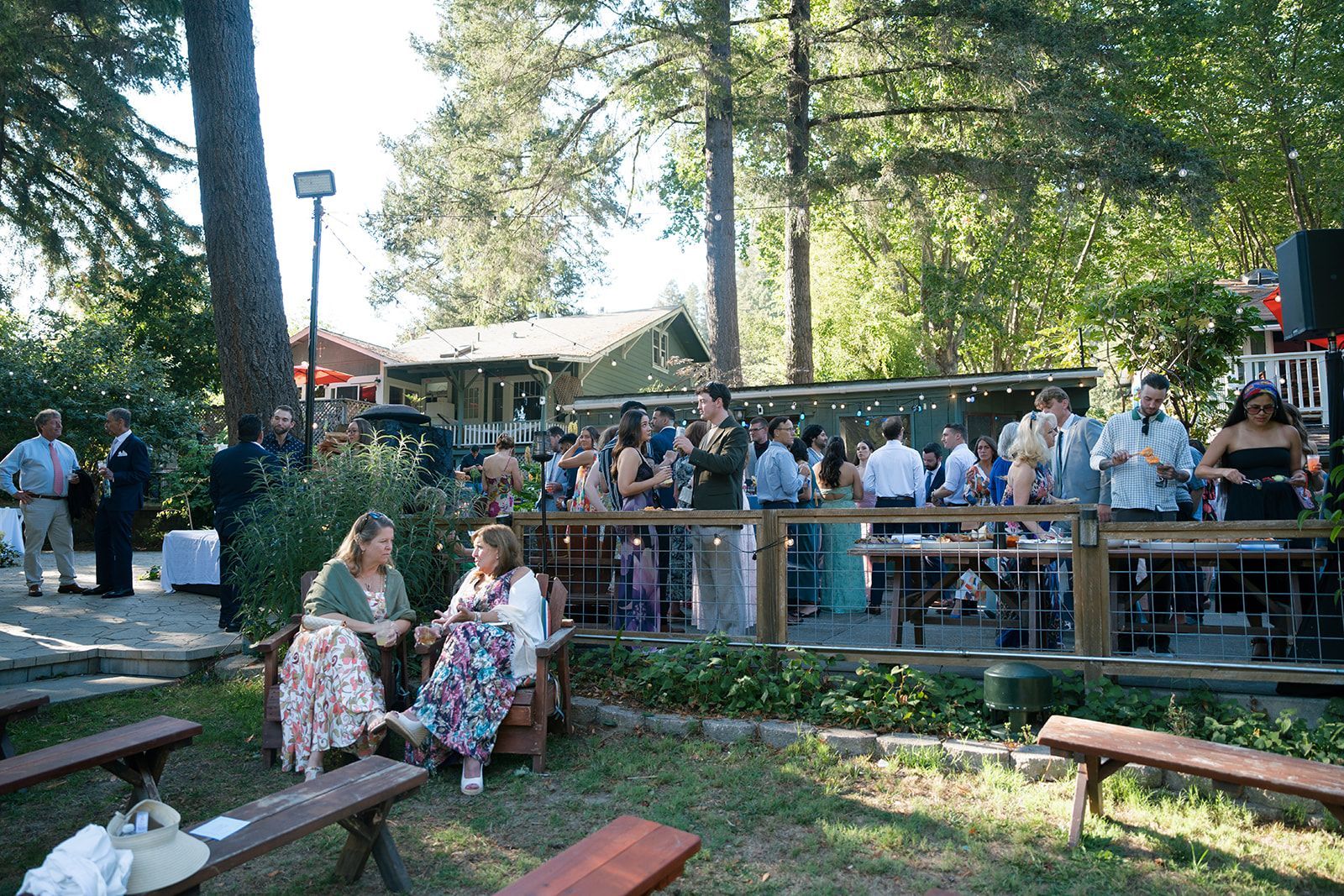 Outdoor party with guests on a deck, some seated on the grass. Trees and a building in the background.
