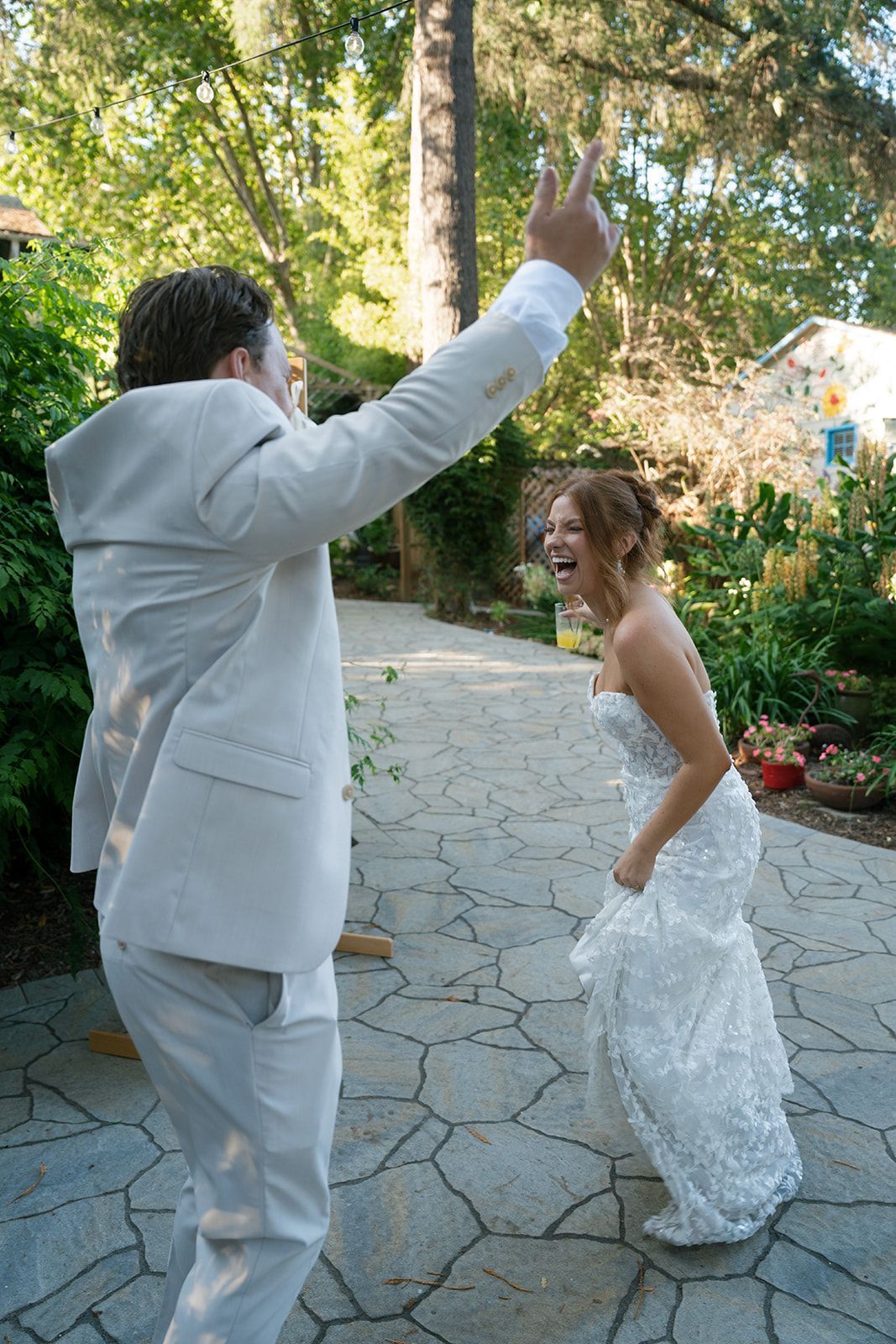 Newlyweds laughing, man in suit cheering, woman in wedding dress, outdoor garden.