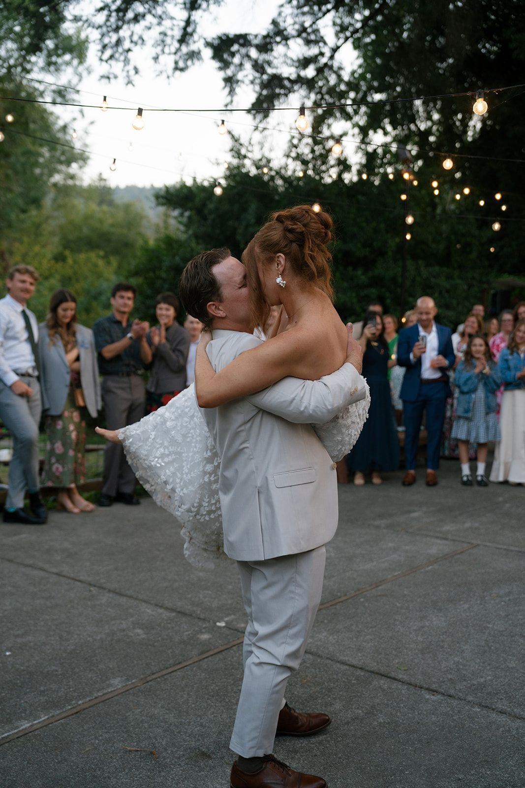 Groom lifts bride, kissing at outdoor wedding. Guests cheer; string lights hang above.
