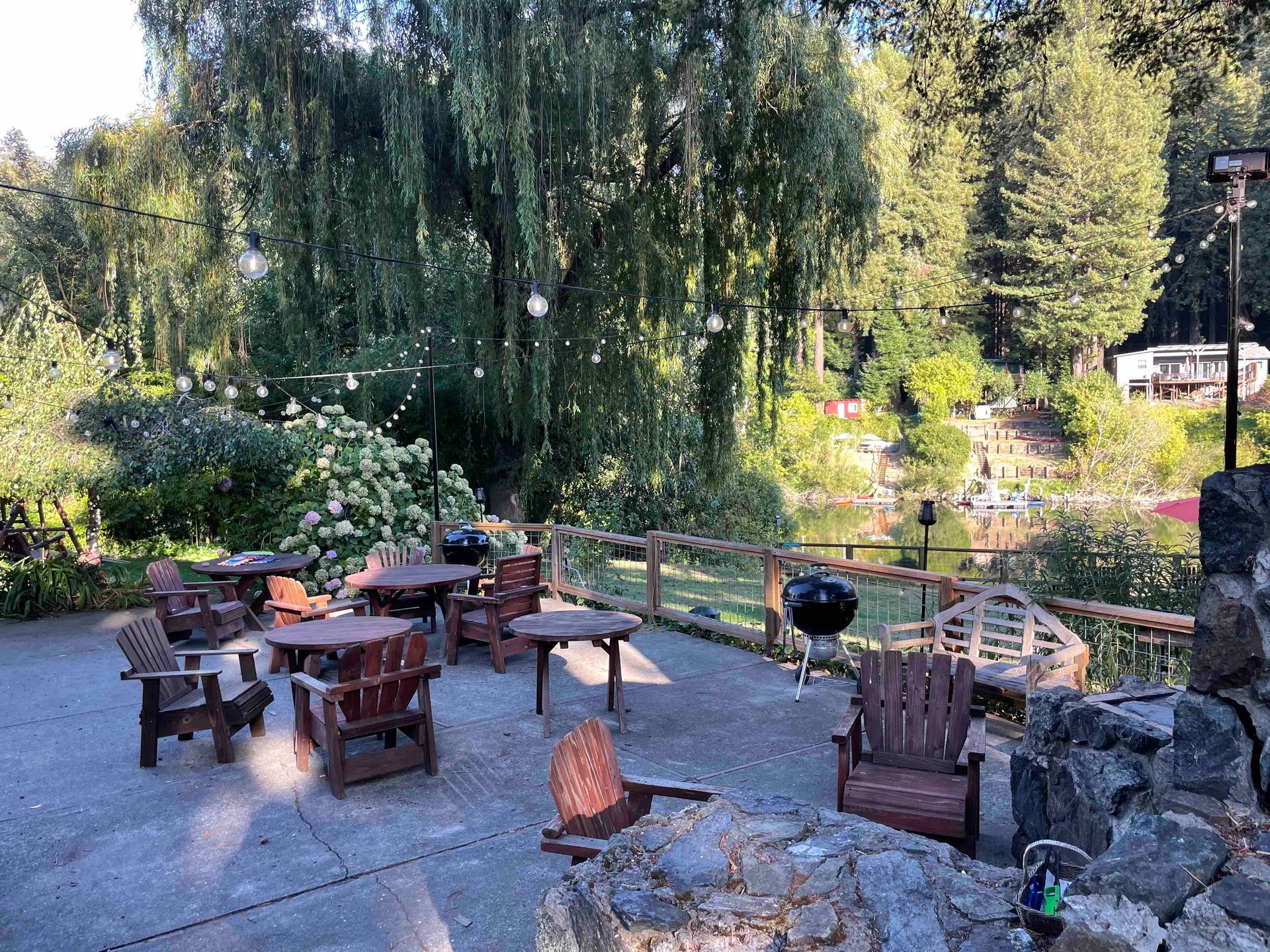 Outdoor patio with wooden furniture, overlooking a lake surrounded by trees. String lights hang above.
