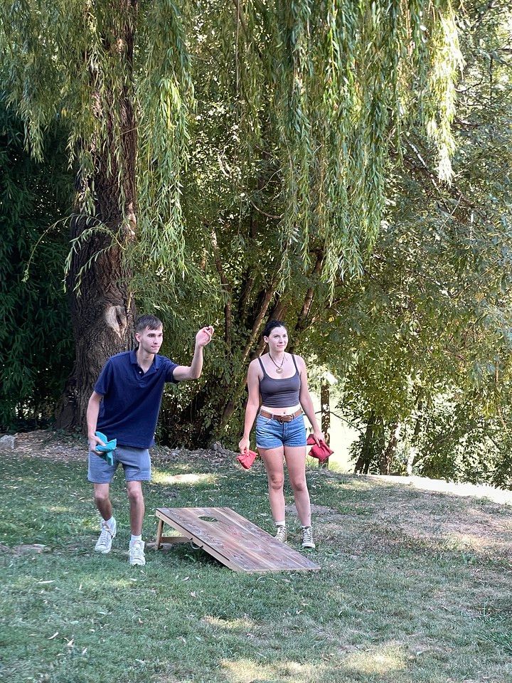 A man throws a bean bag at a cornhole board, a woman watches in a grassy outdoor setting.