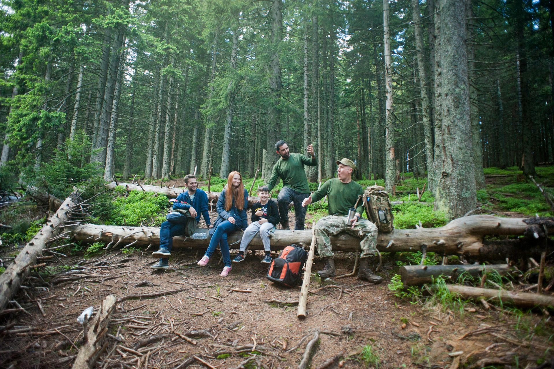 Group of hikers resting on logs in a forest clearing, surrounded by tall green trees.