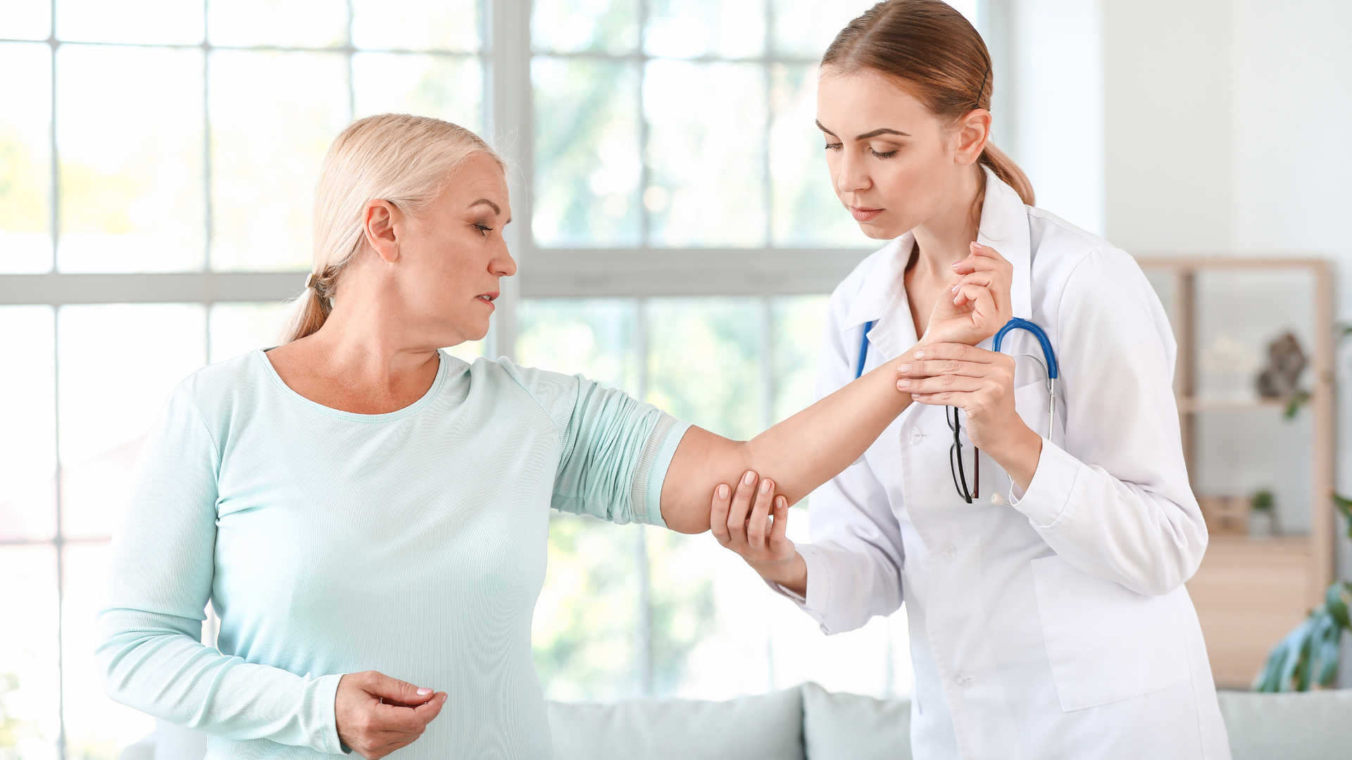 A doctor examines an older woman's arm in a well-lit room. The doctor wears a white coat and stethoscope. The patient appears concerned.