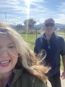 Woman smiling, taking a selfie with a man wearing sunglasses outdoors on a sunny day.
