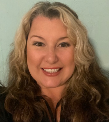 Woman with long, wavy brown and blonde hair, smiling. Wearing a black shirt, against a light blue background.