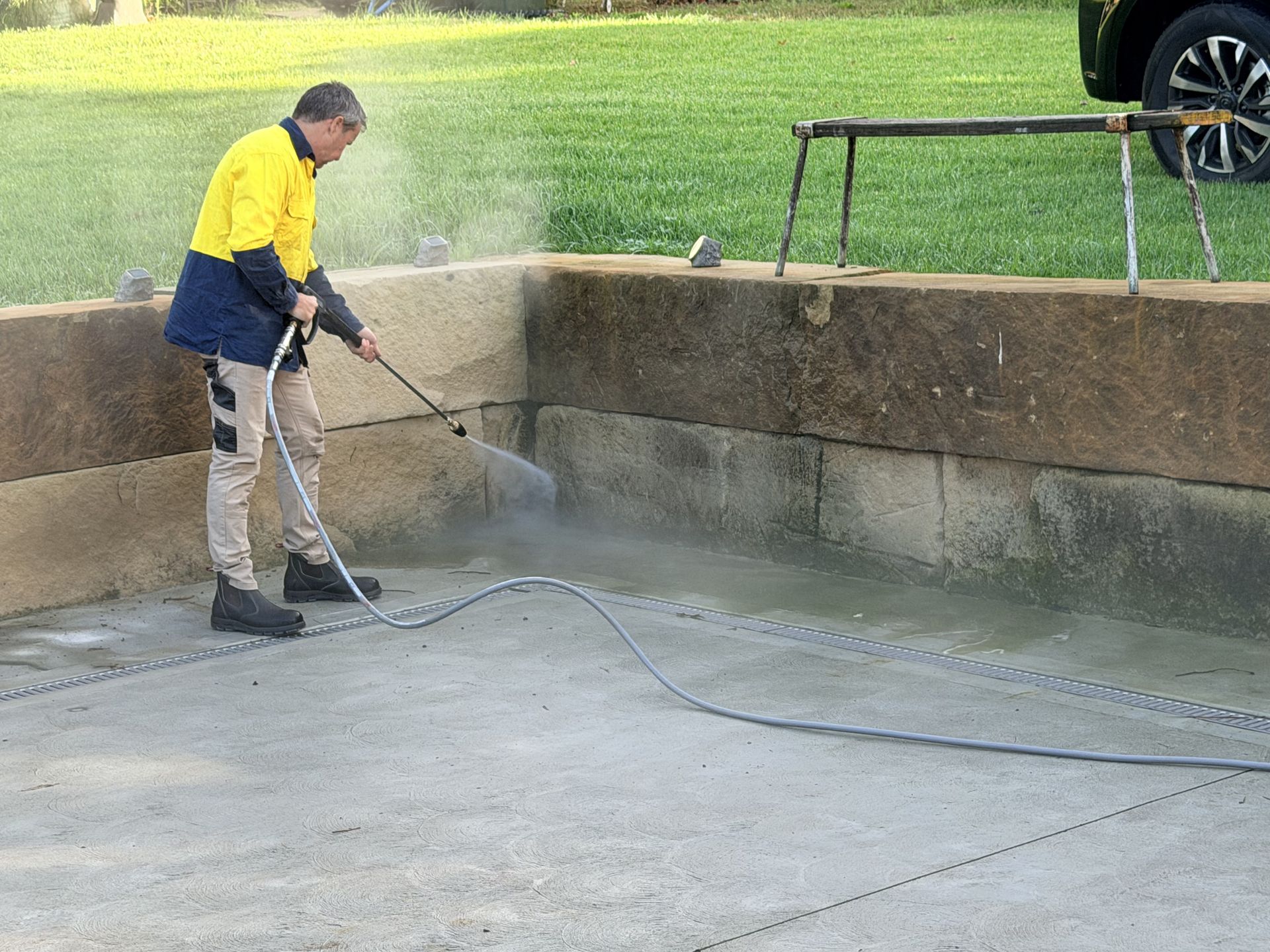 Worker in yellow jacket pressure-washing a concrete area beside a retaining wall.