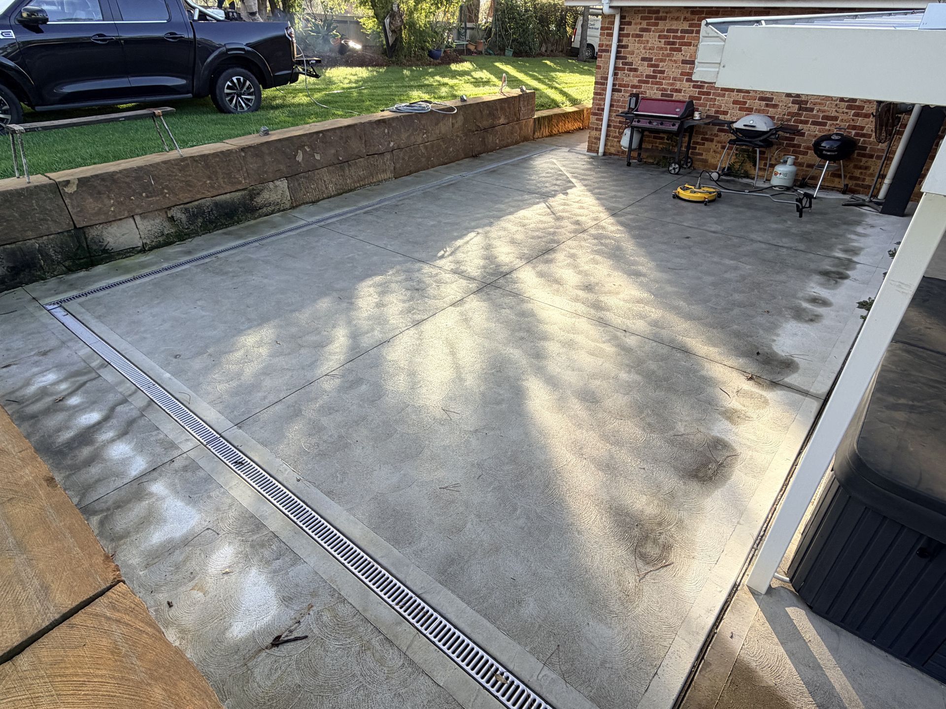 Patio concrete slab with drainage grates beside a brick house, in a backyard driveway area.