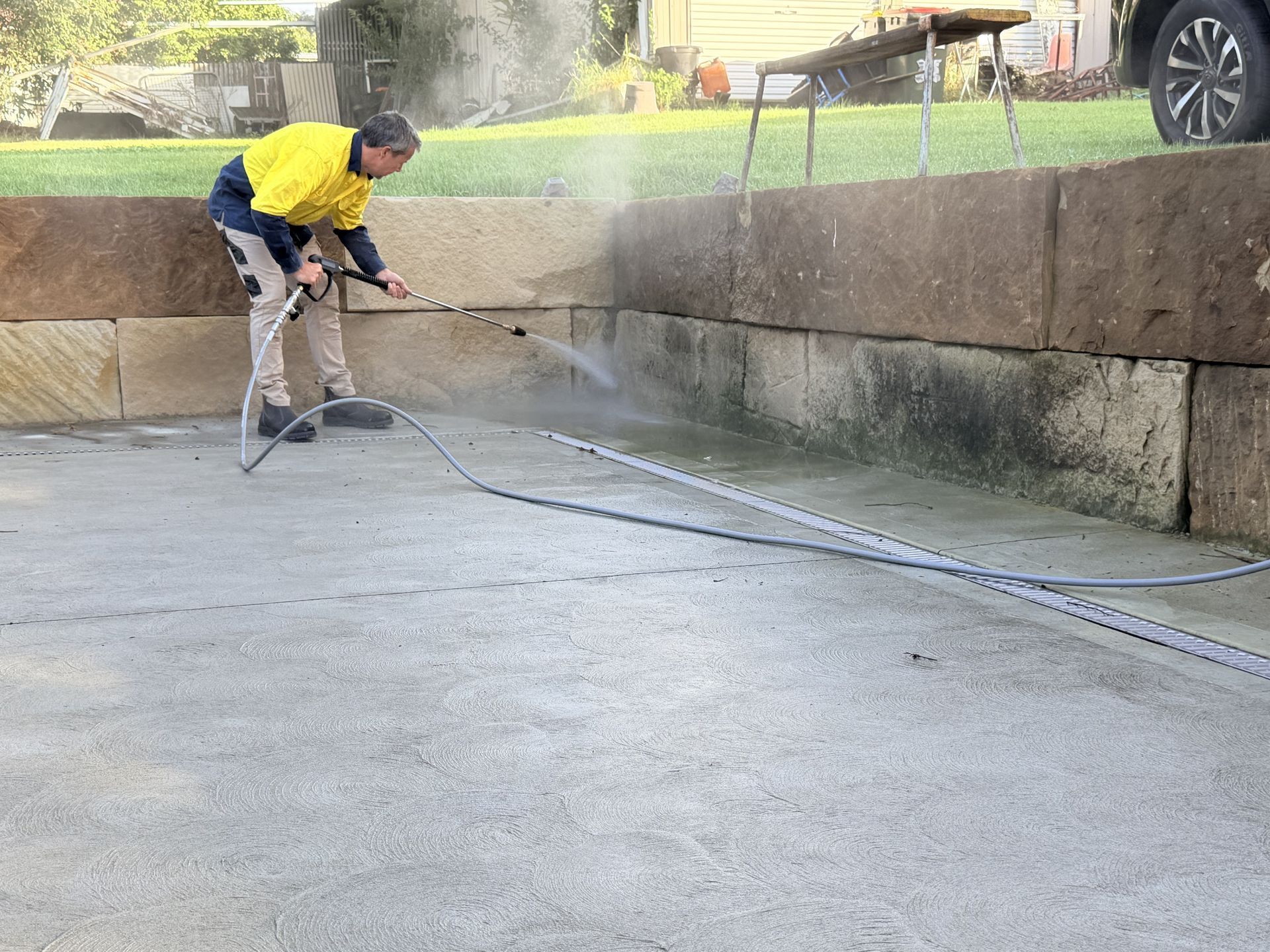Worker in yellow shirt pressure-washing a concrete sidewalk beside a stone wall and fountain