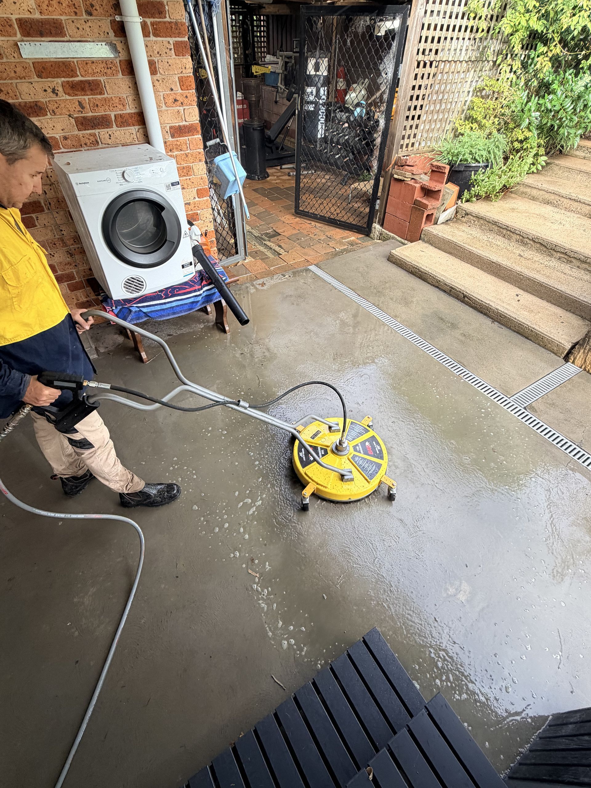 Person using a yellow pressure washer on a wet concrete patio beside a brick house