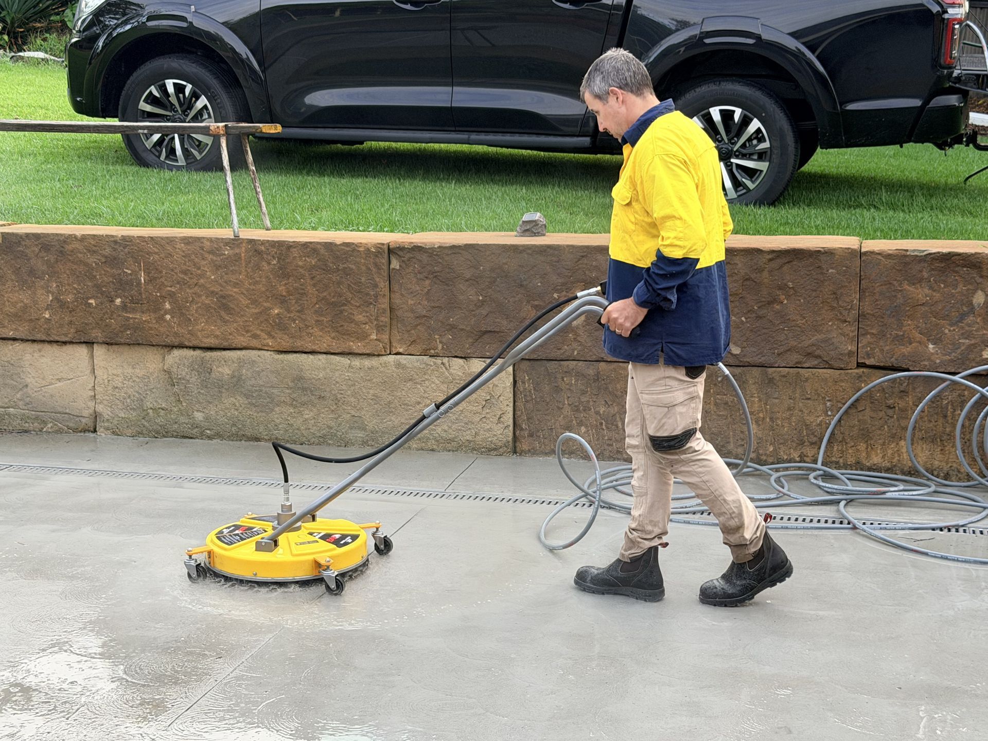 Worker using a yellow floor-cleaning machine on a sidewalk beside a parked black SUV