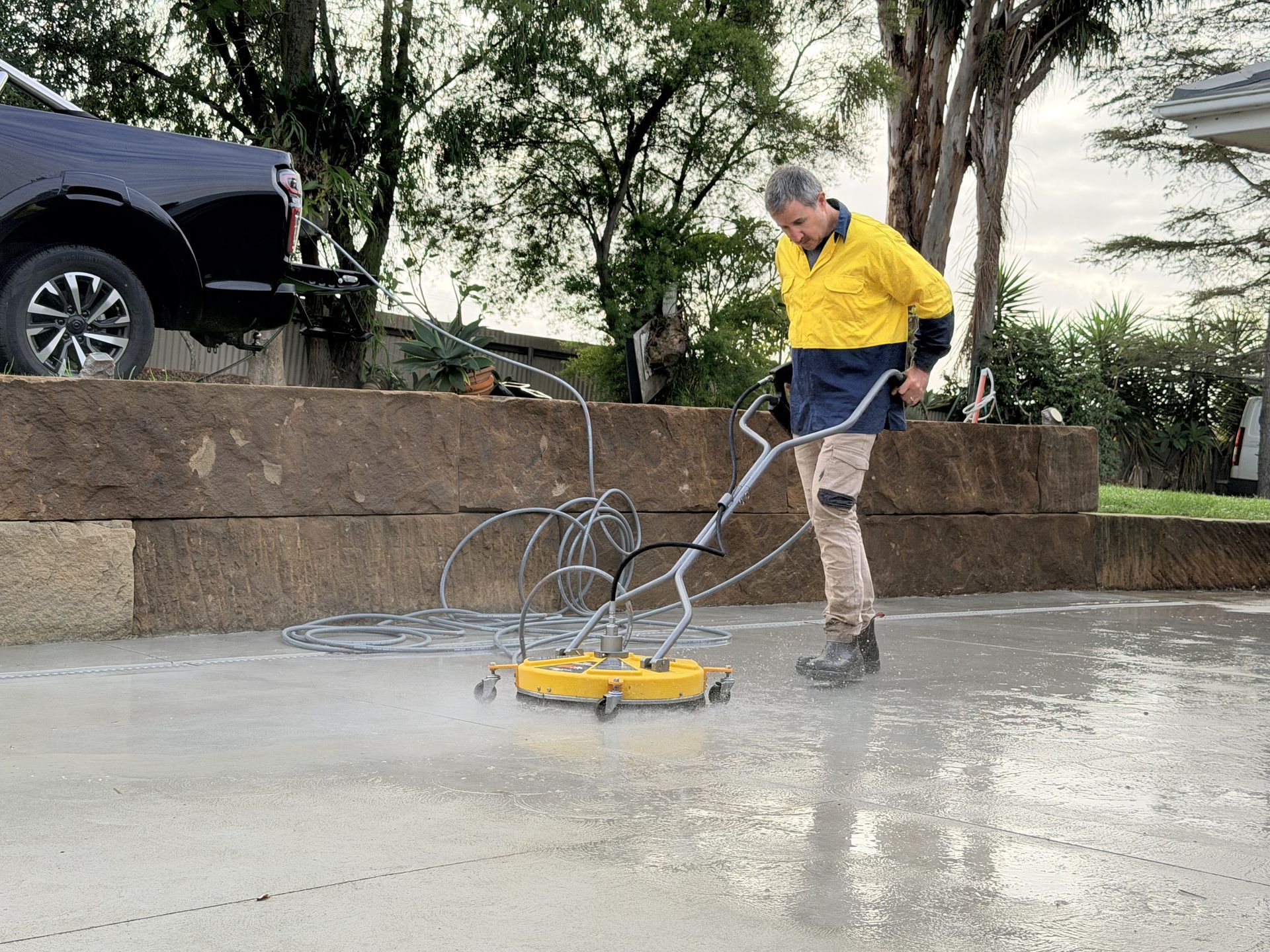Worker pressure-washing a driveway with a yellow surface cleaner beside a stone wall and parked SUV