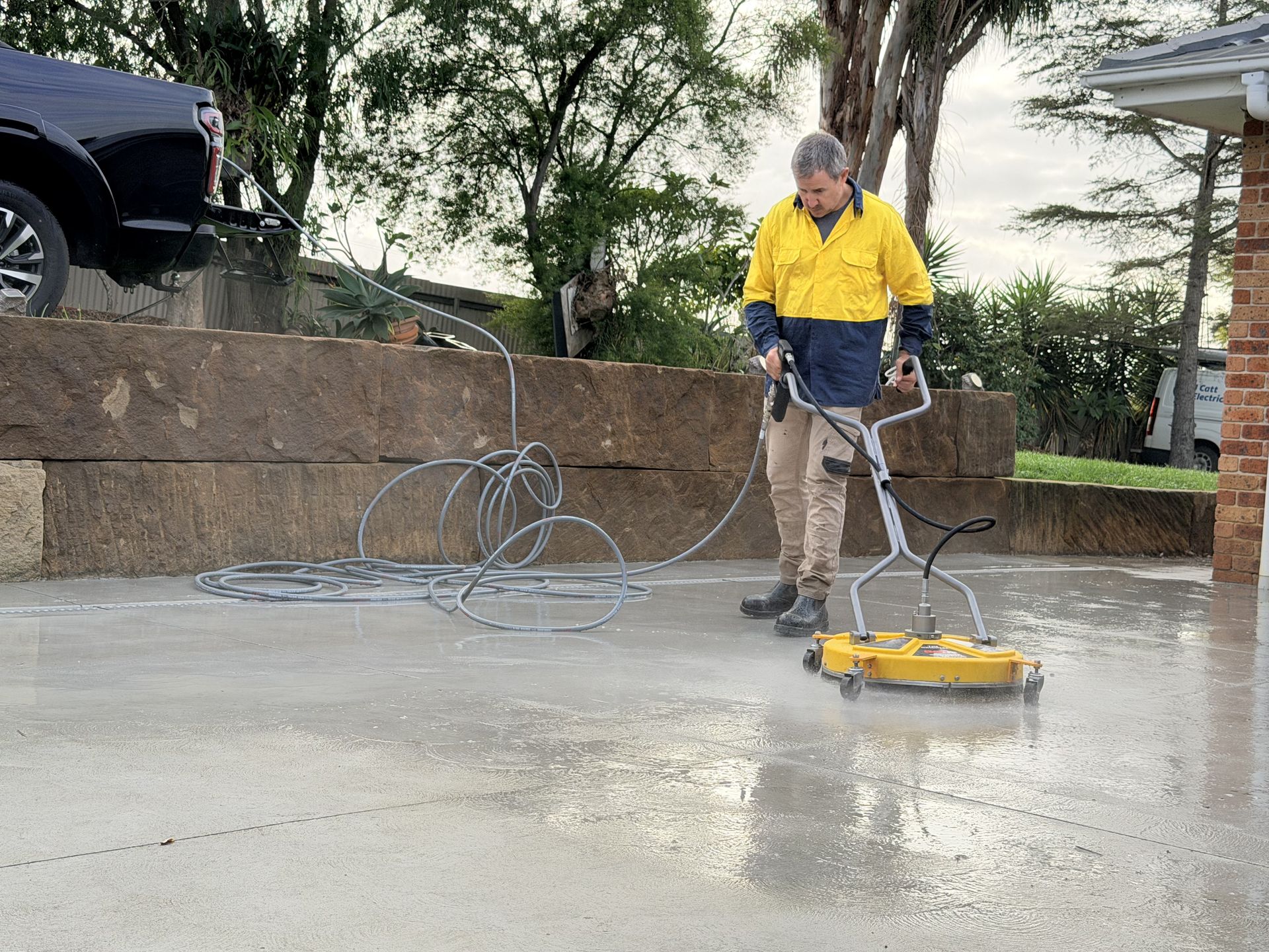 Person pressure-washing a wet driveway with a yellow cleaning machine, hose coiled nearby.