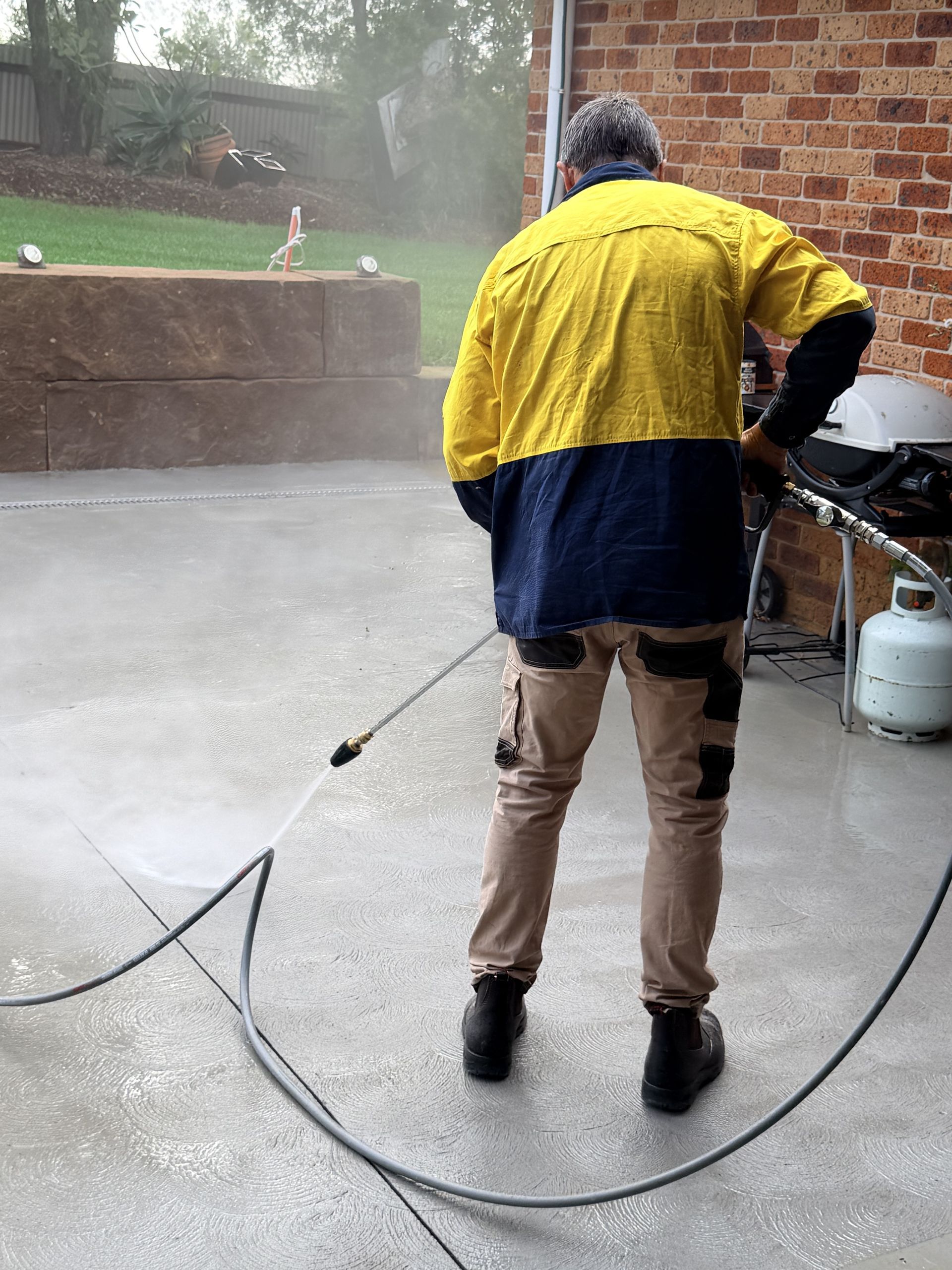 Person pressure washing a wet concrete driveway beside a patio, wearing a yellow and navy jacket.