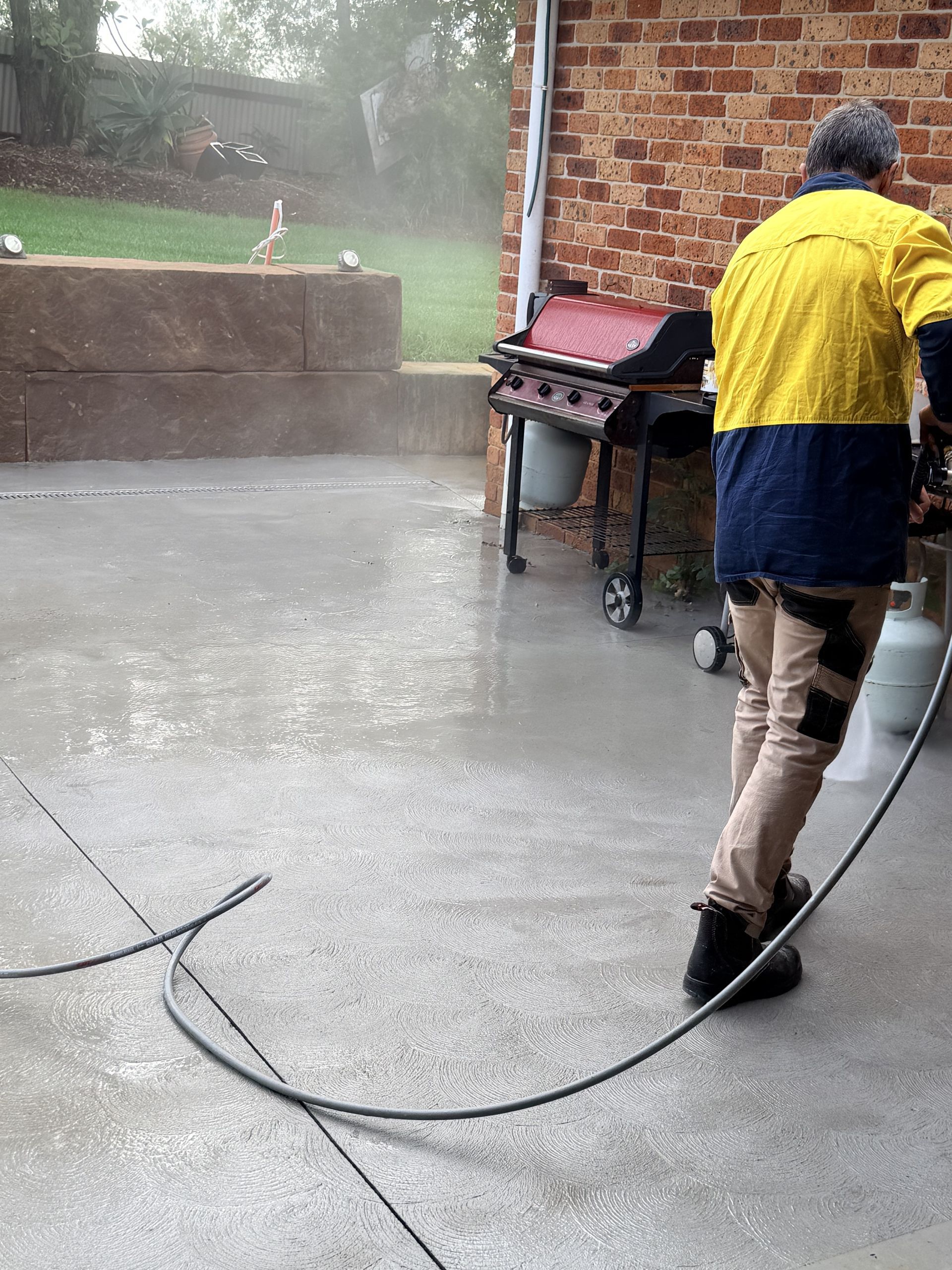 Person pressure-washing a concrete patio beside a brick wall and grill in the rain