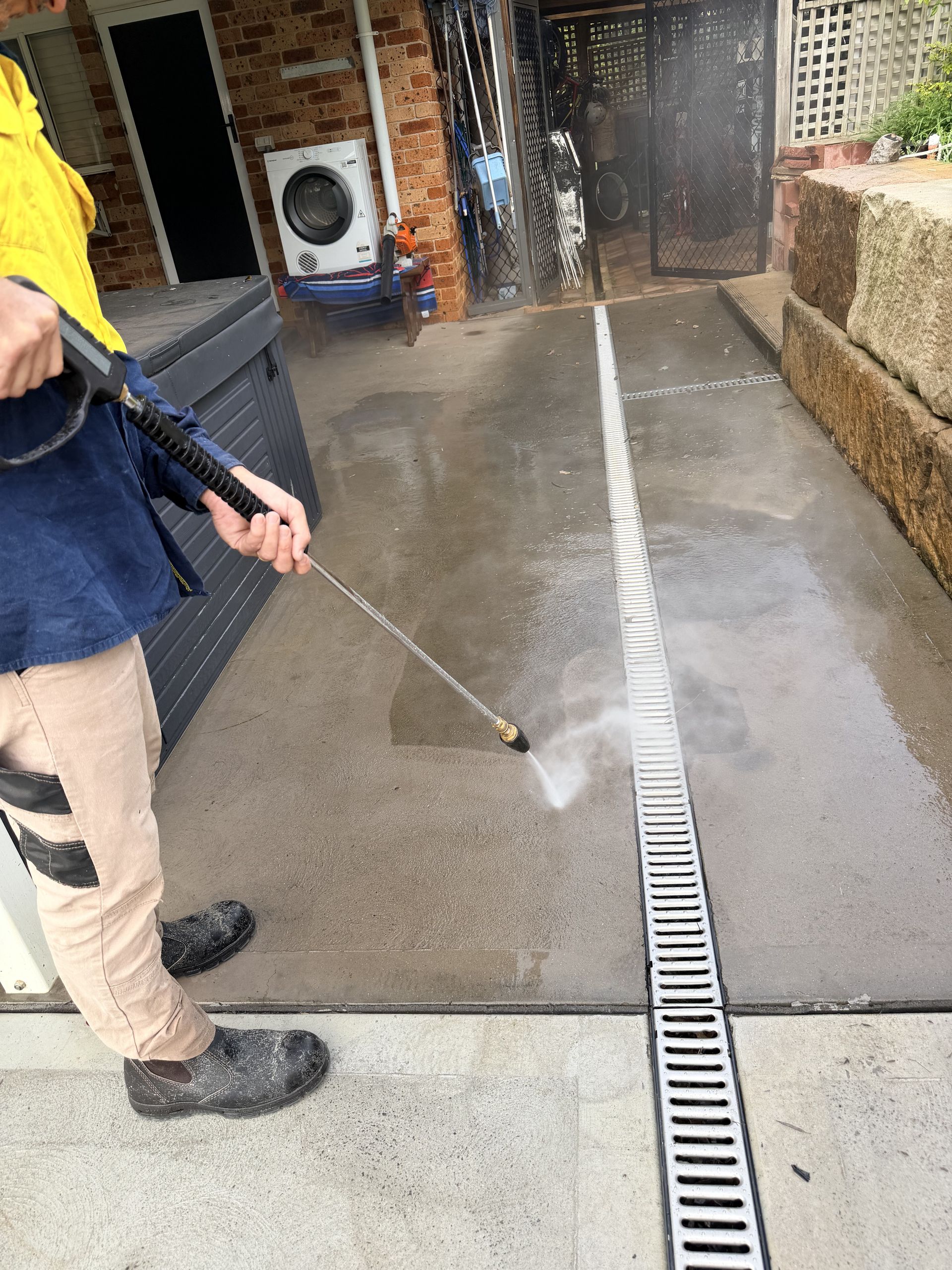 Person pressure-washing a wet concrete walkway beside a building and drainage channel.
