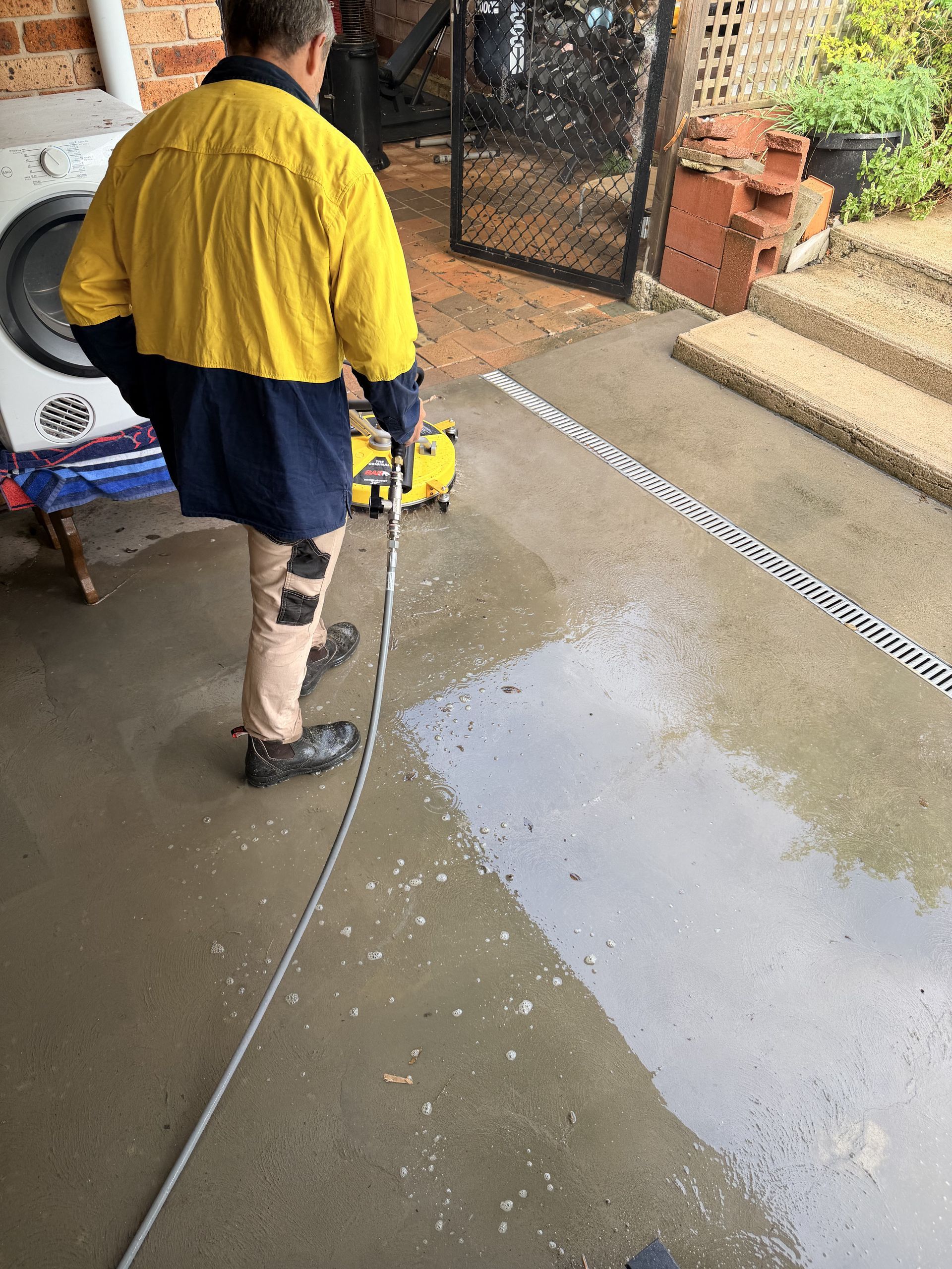 Worker pressure-washing a concrete floor in a garage, wearing a yellow jacket and blue pants.