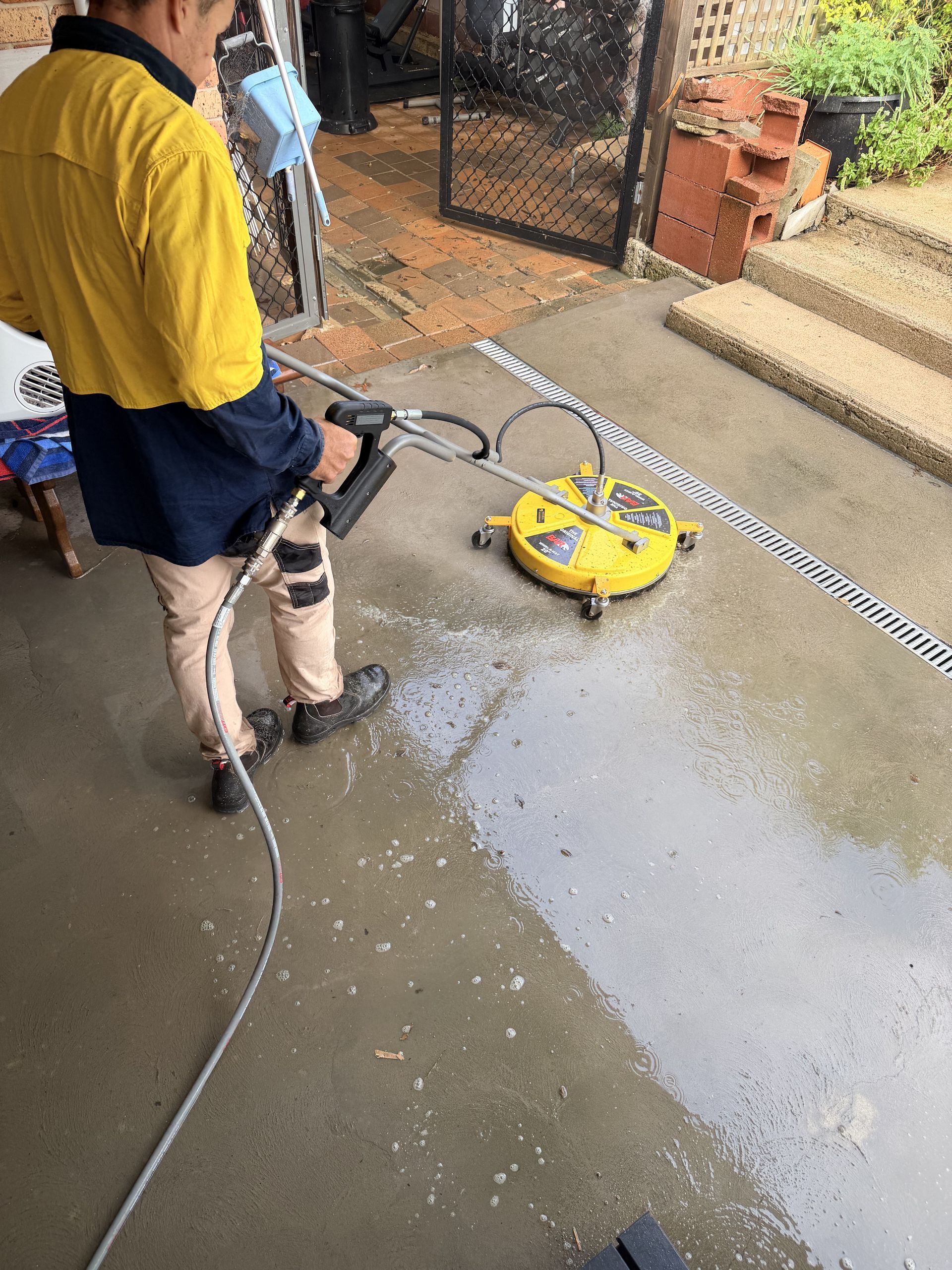 Person using a pressure washer to clean a wet concrete patio near a yellow machine and gate.