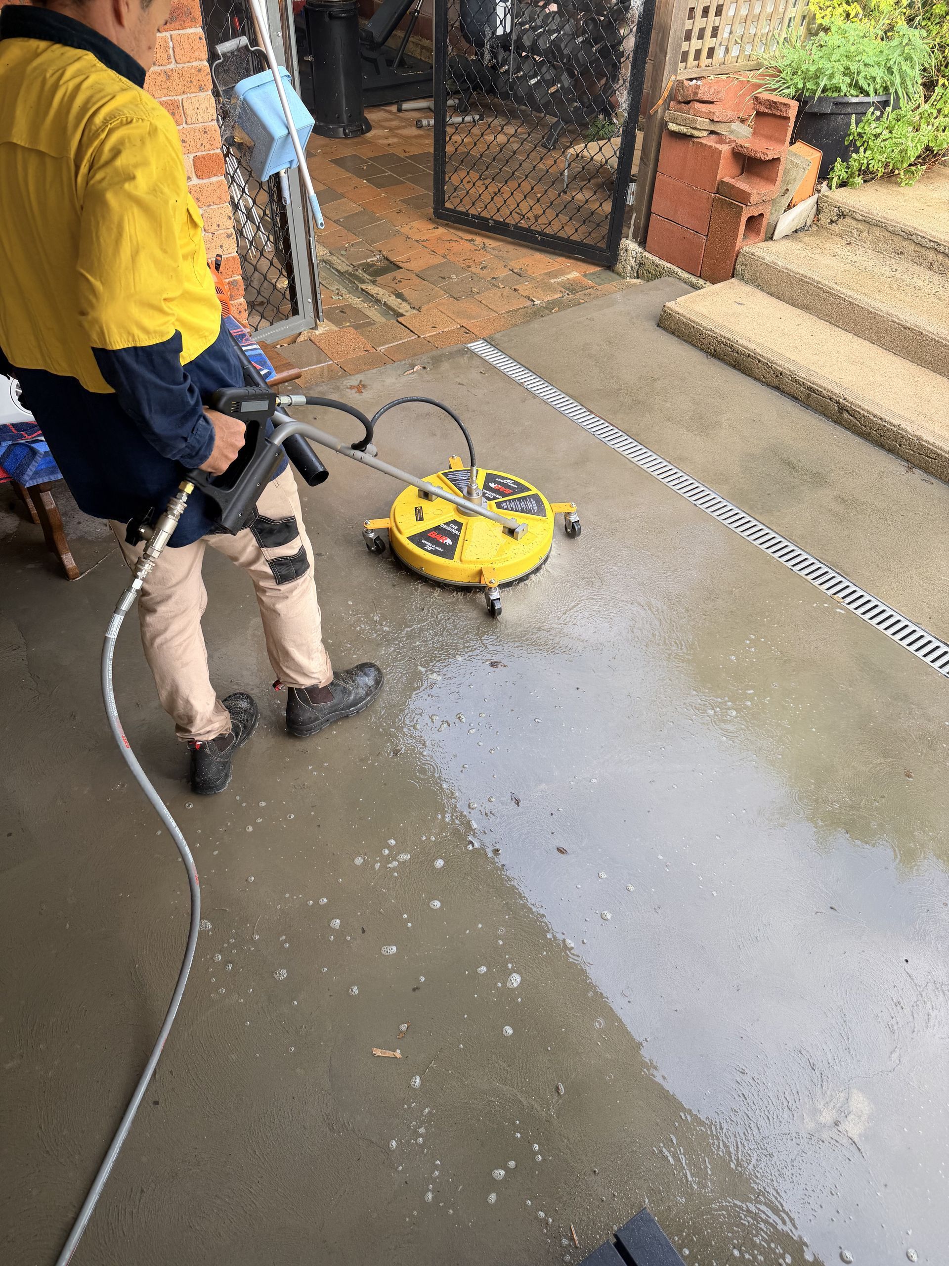 Person pressure-washing a wet concrete driveway with a yellow cleaner near a brick gate and steps