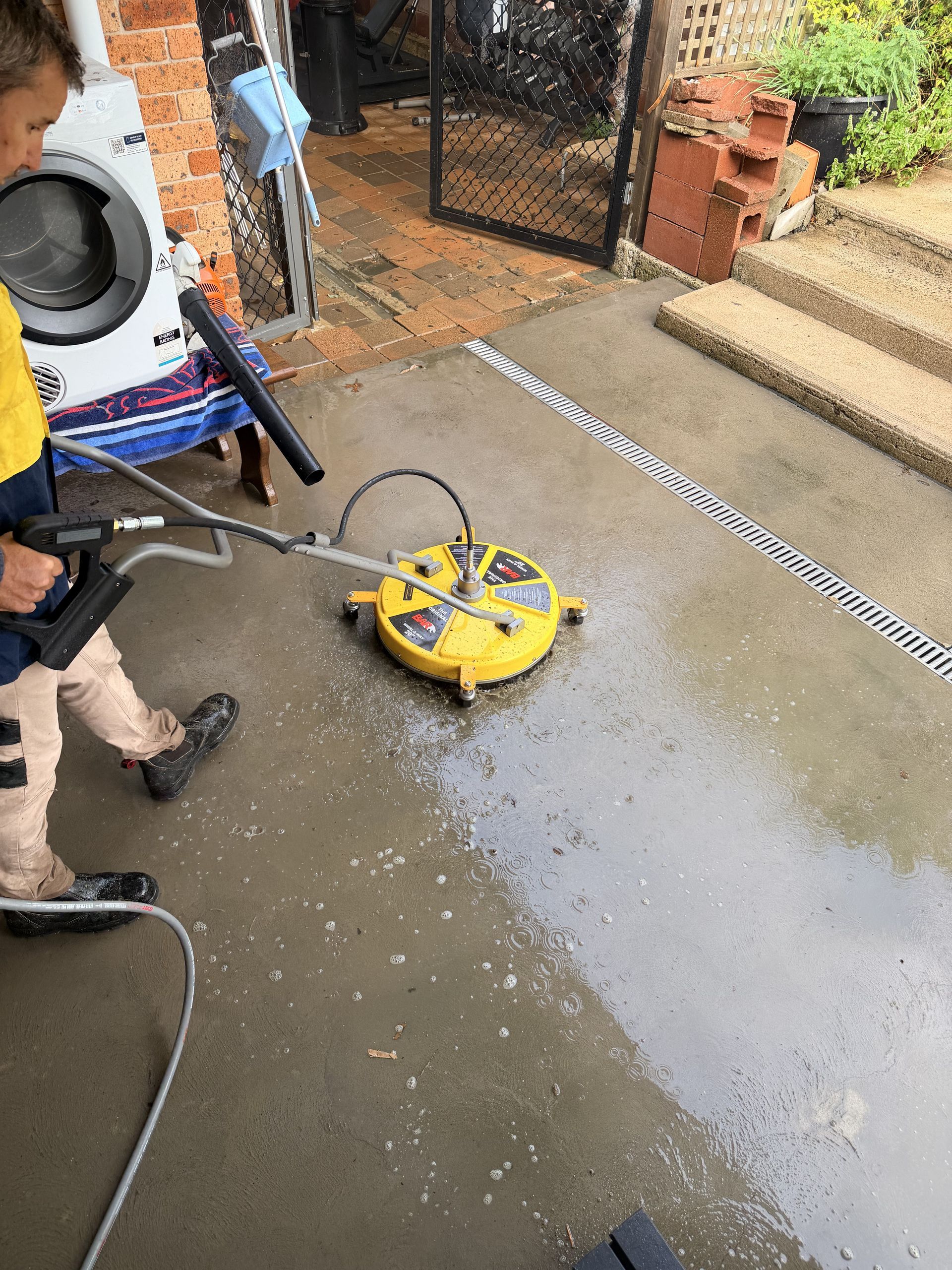 Person pressure-washes a wet driveway with a yellow cleaner near steps and a gate.