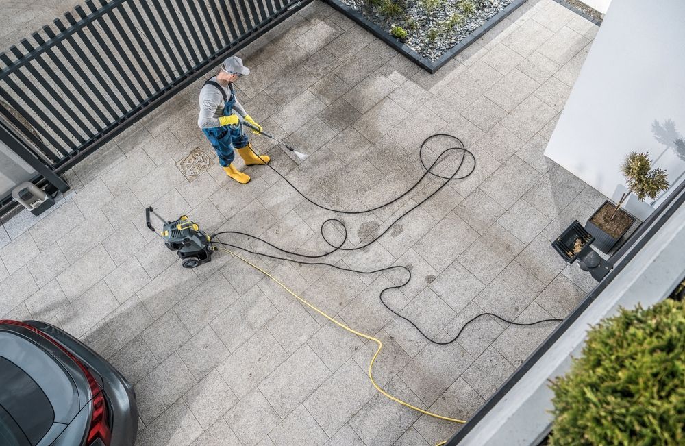 Person pressure-washing a driveway beside a black gate, with hoses and a red car nearby.