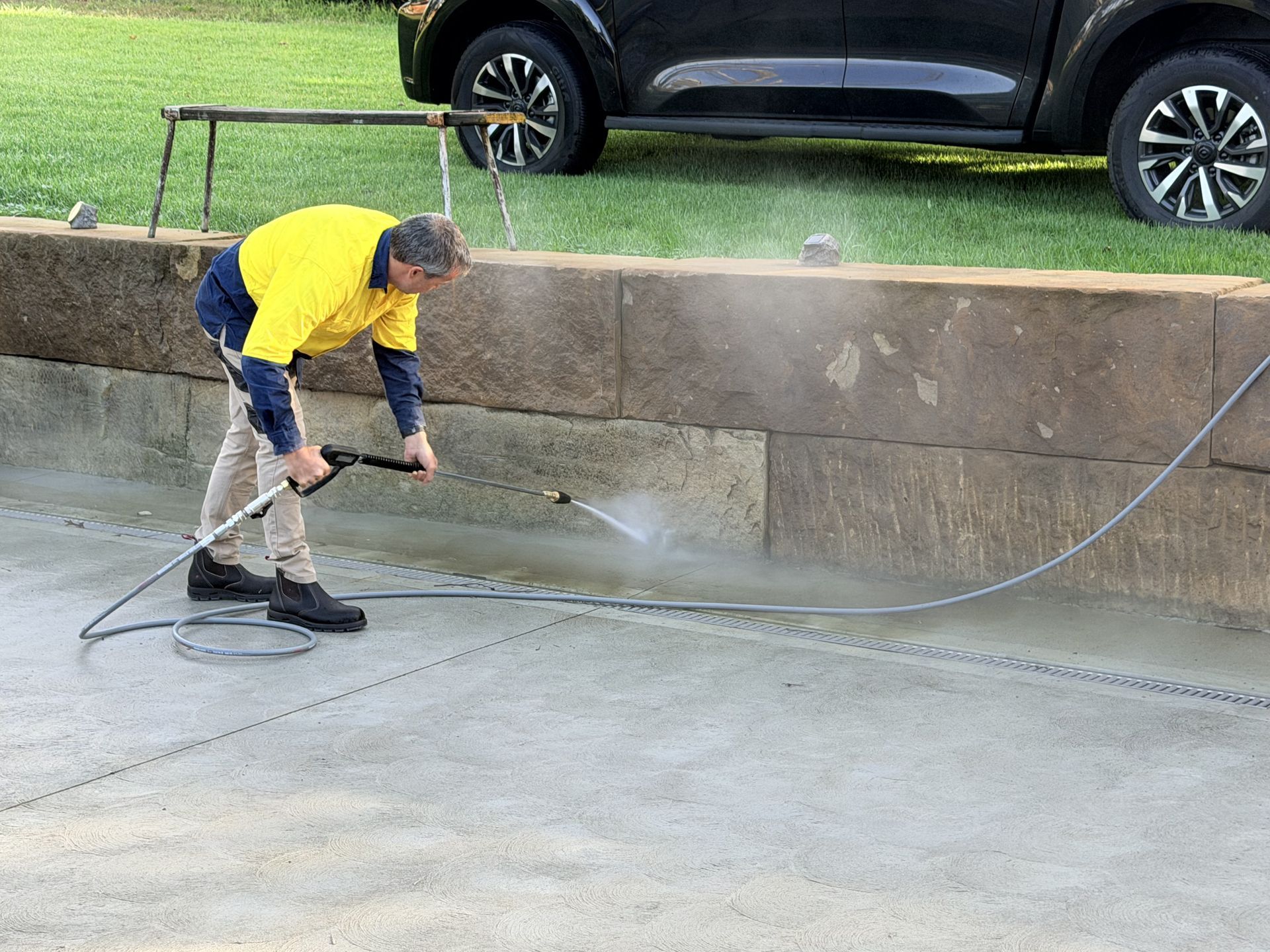 Worker in a yellow shirt pressure-washing a concrete driveway beside a parked SUV.