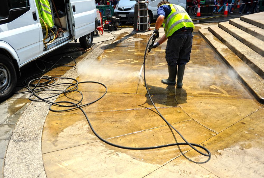 Worker pressure-washing a wet concrete area beside parked vans, with hoses spread across the ground