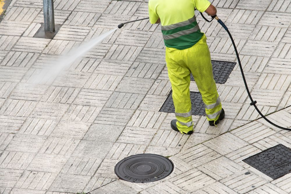 Worker in neon yellow uniform cleaning pavement with a pressure washer on a tiled street.
