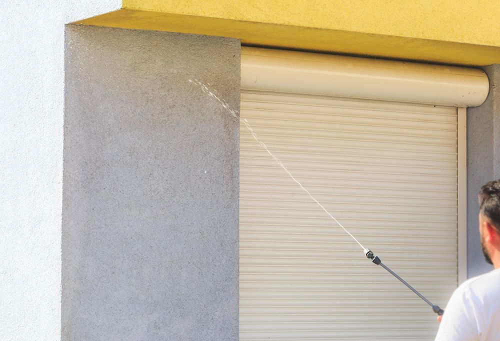 Person washing a roll-up shutter with a pressure washer next to a stucco wall