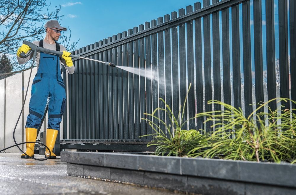 Worker pressure-washing a black metal fence beside a curb, wearing blue overalls and yellow gloves and boots