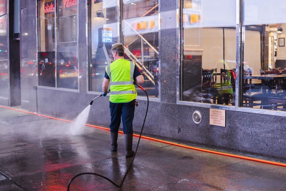 Worker in neon safety gear cleaning a sidewalk beside a street with manhole covers and traffic cones