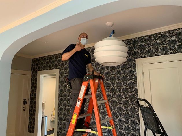 A man wearing a mask is standing on a ladder fixing a light fixture.