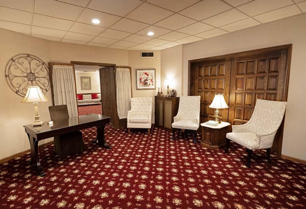 A waiting room with red carpet, chairs, lamps, and a dark desk. Wooden doors lead to other rooms.