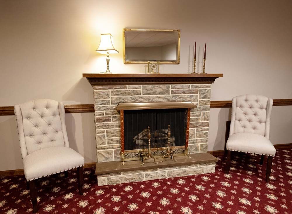 Fireplace flanked by two button-tufted chairs; a lamp and mirror adorn the mantel. Red patterned carpet.
